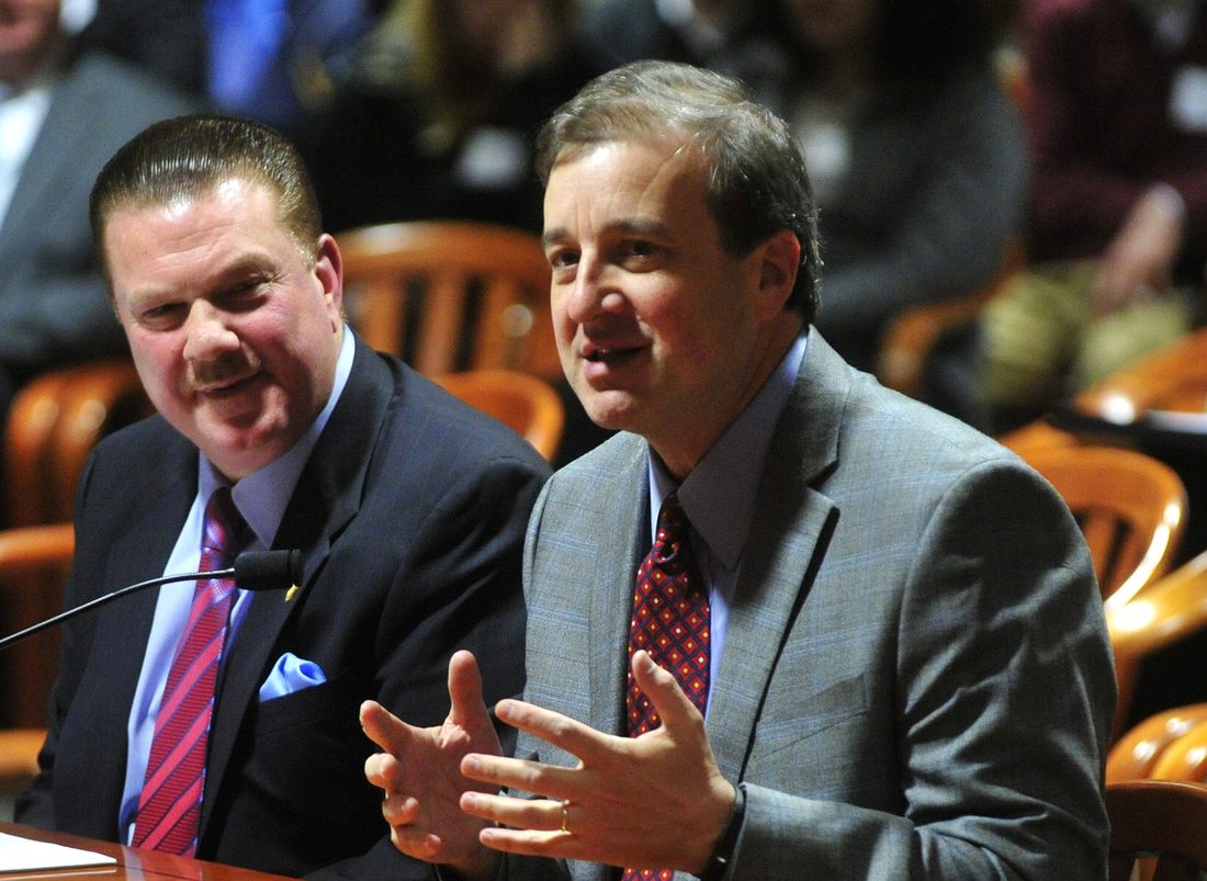 Scott Yates testifying at a Michigan legislative hearing, gesturing as he speaks, while the bill's sponsor looks on and smiles.
